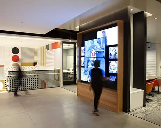 Modern interior: large digital display. Two people, one interacts. Geometric patterns, colorful walls, seating visible behind.